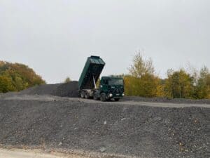 Ein grüner Lastwagen kippt Kies auf einem grauen Hügel, umgeben von Herbstbäumen.