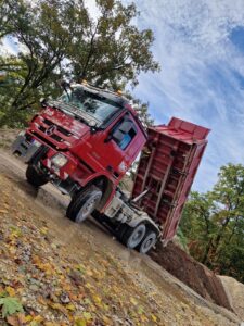 Roter Lkw kippt Erde auf Baustelle ab, Herbstblätter im Vordergrund, bewölkter Himmel im Hintergrund.