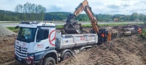 Lastwagen und Bagger auf Baustelle, Arbeiter in orangefarbener Kleidung, bewaldete Landschaft.