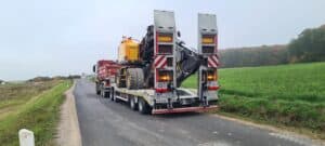 Ein Lkw transportiert einen großen gelben Bagger auf einer Landstraße in ländlicher Umgebung.