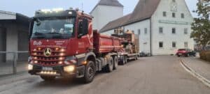 Roter Lastwagen mit Anhänger auf Straße, Gebäude im Hintergrund, Baum rechts.